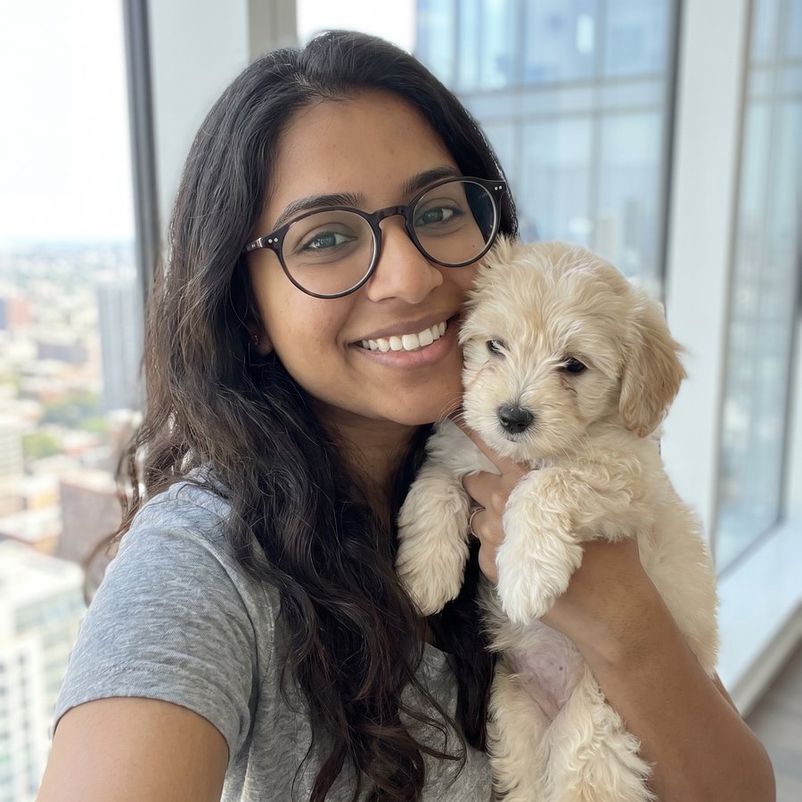 A smiling woman with long dark hair kneels on the floor looking at a golden retriever puppy.